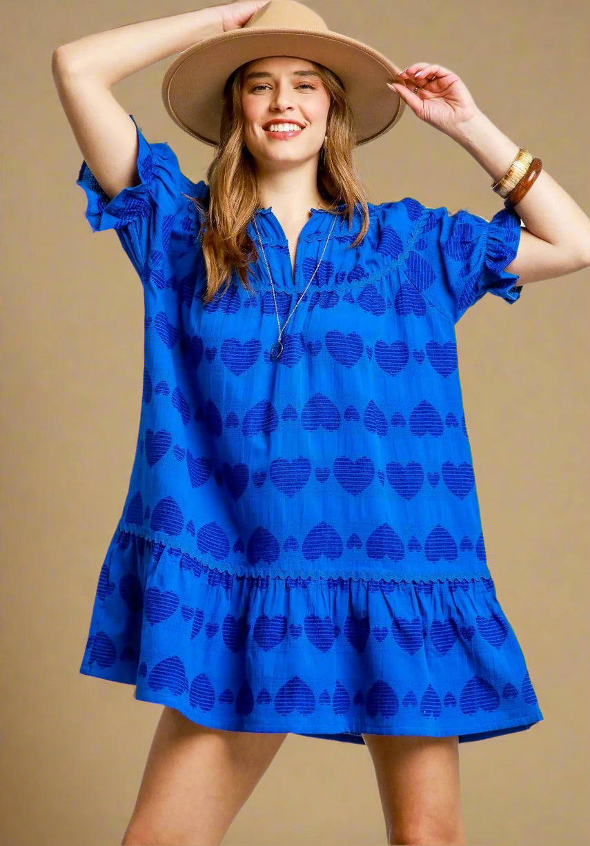 Woman wearing a blue dress with heart pattern and a wide-brimmed hat on a light gray background