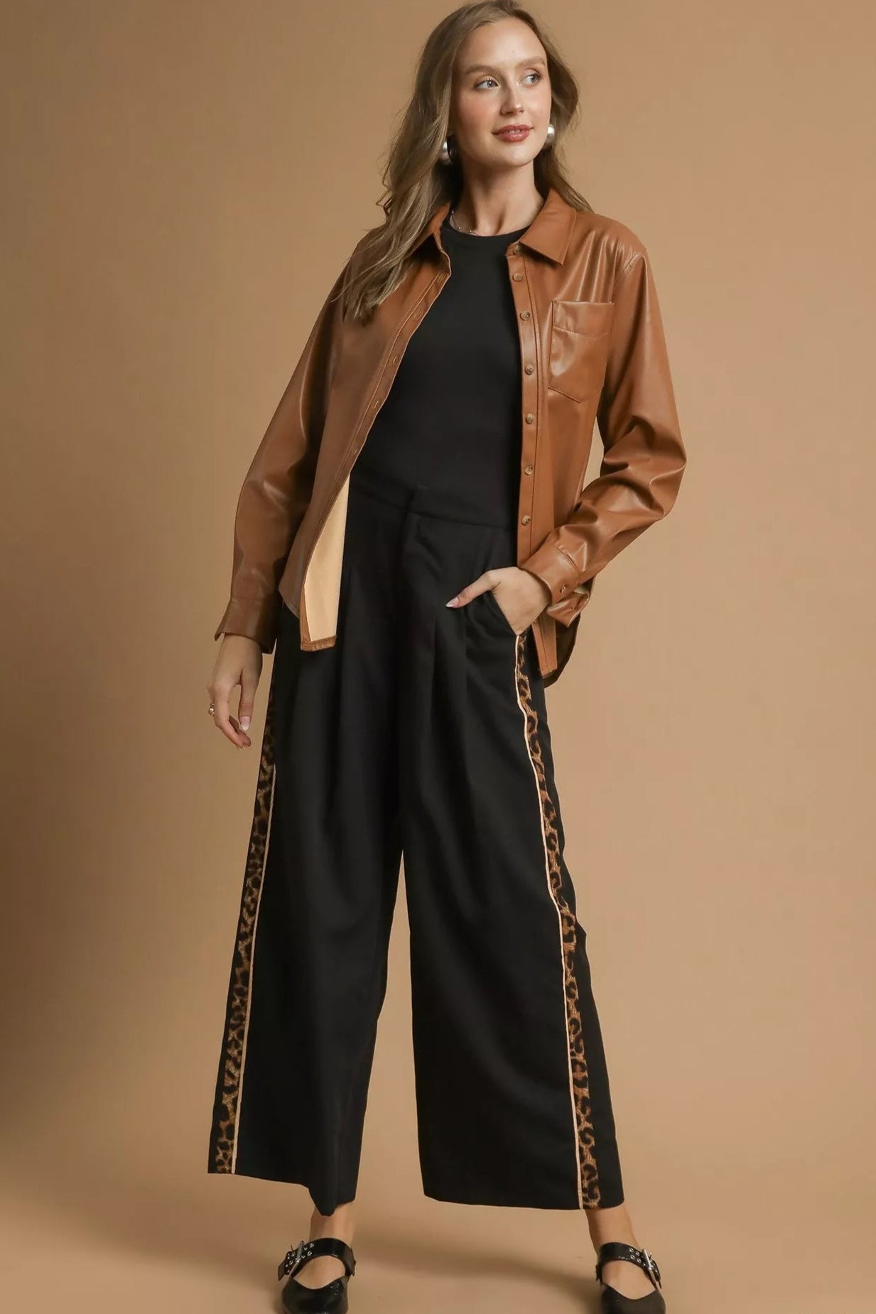 Woman wearing a brown leather jacket and black pants with leopard print trim on a beige background