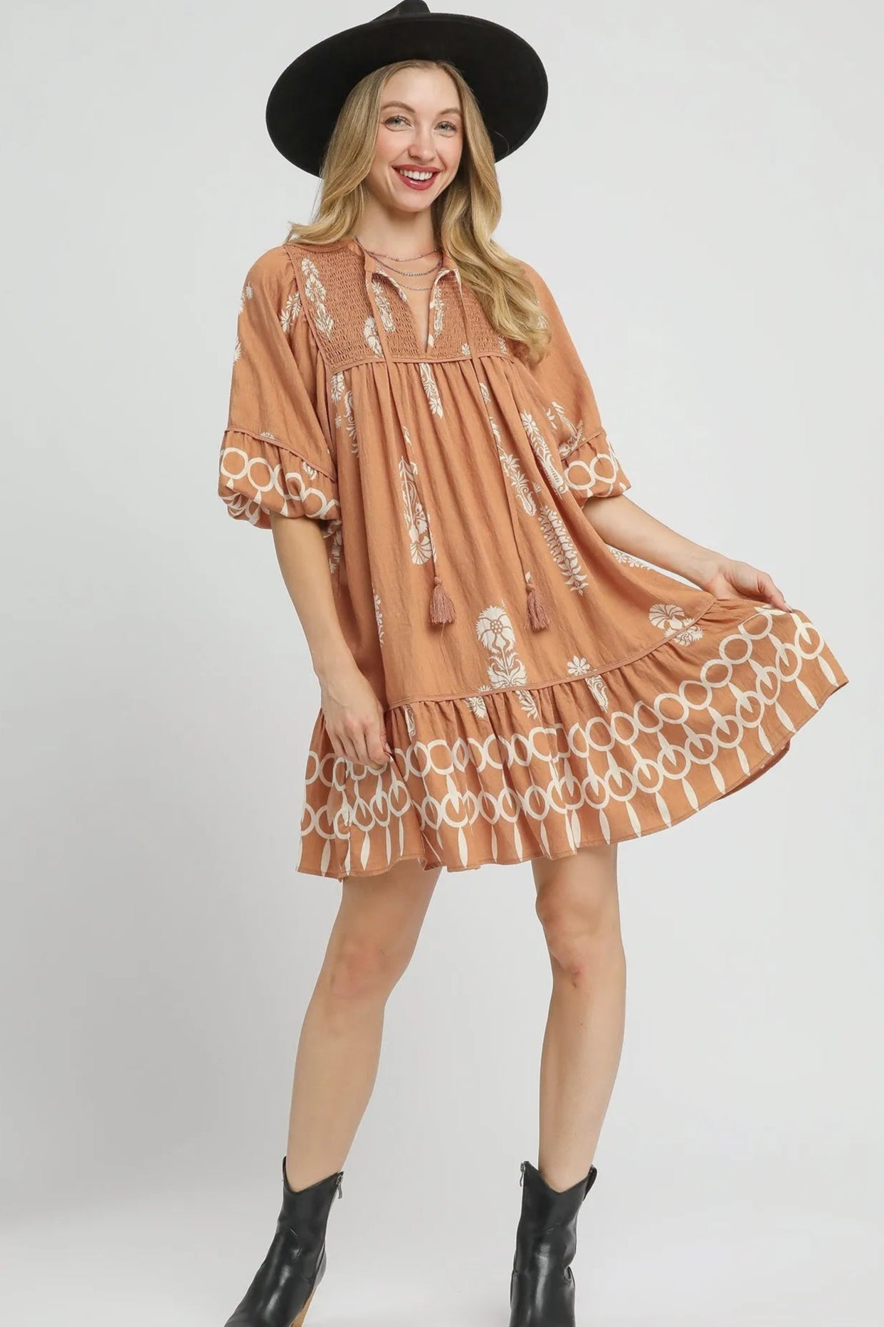 Woman wearing a brown dress with white patterns, black boots, and a black hat on a white background.