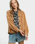 A woman modeling a tan button-up overshirt with gathered sleeves, worn over a black and white leopard print top and paired with denim shorts, against a light gray studio background.