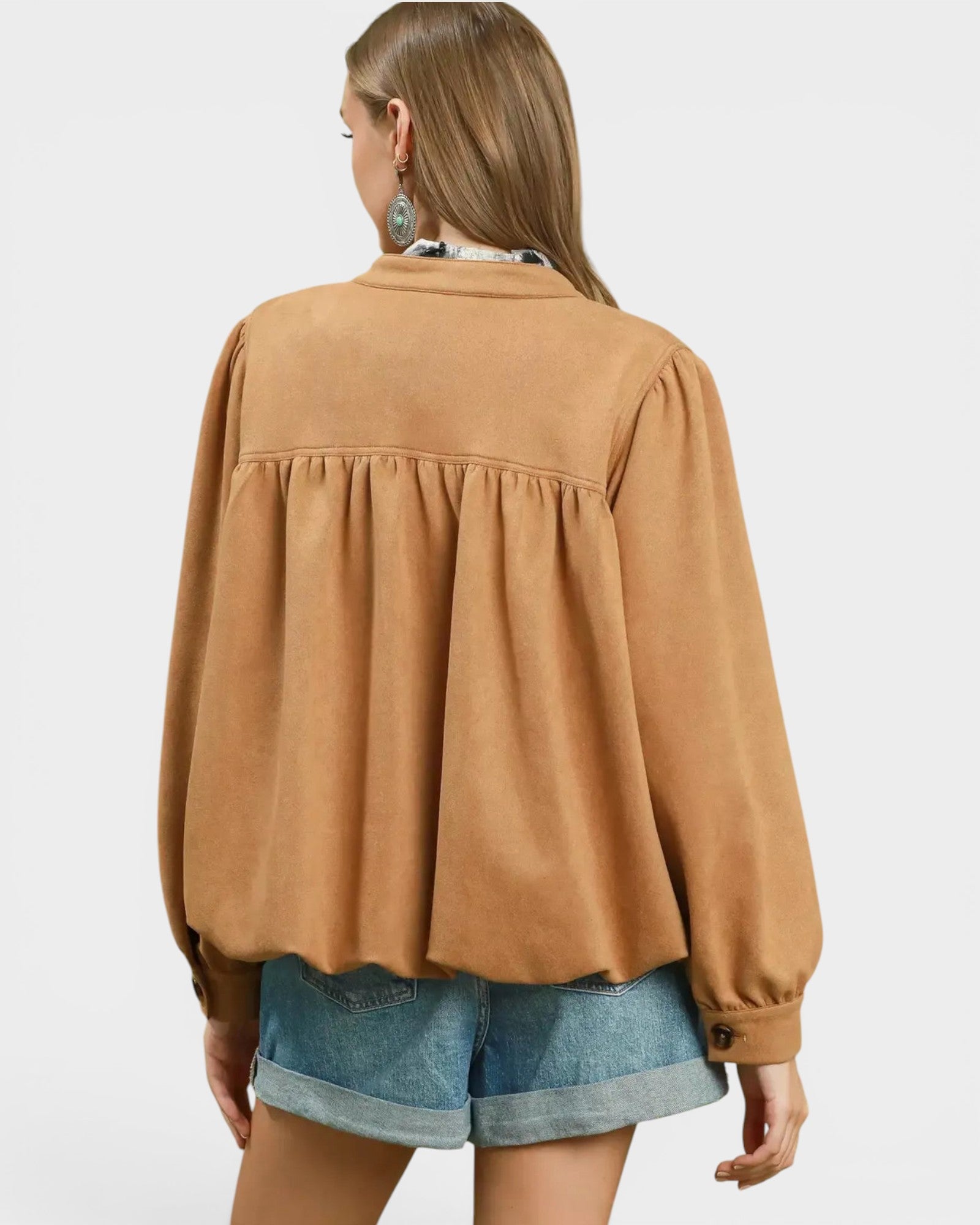 A woman shown from the back wearing a camel-colored long-sleeve blouse with a gathered peplum-style back seam and denim shorts, against a light gray studio background.