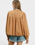 A woman shown from the back wearing a camel-colored long-sleeve blouse with a gathered peplum-style back seam and denim shorts, against a light gray studio background.