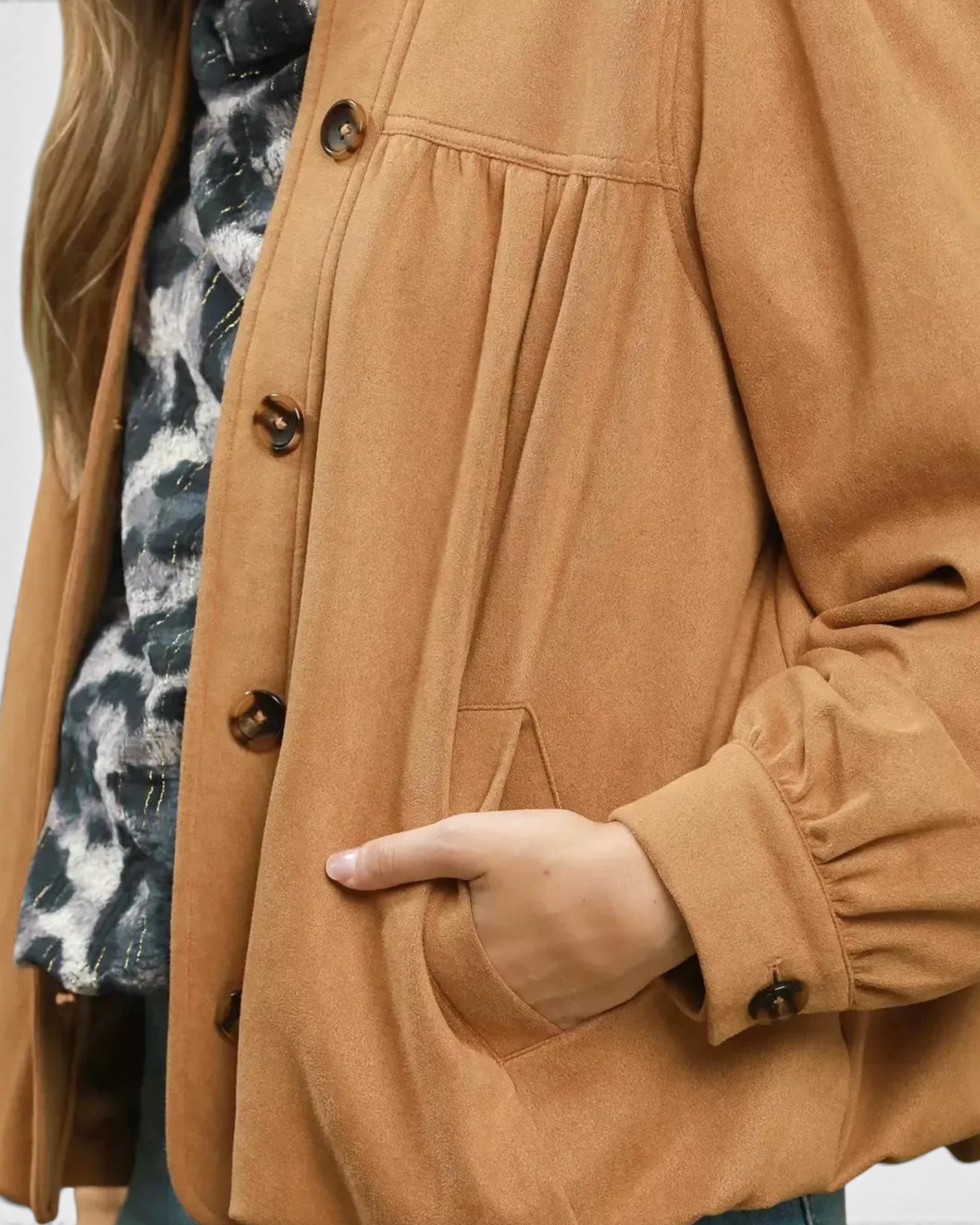 A close-up of a person wearing a camel brown button-up jacket with gathered sleeves and a hand tucked into a pocket, layered over a black and white tie-dye top.