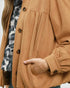A close-up of a person wearing a camel brown button-up jacket with gathered sleeves and a hand tucked into a pocket, layered over a black and white tie-dye top.