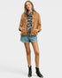 A woman modeling a tan utility jacket over a black and white patterned top, paired with denim shorts and black ankle boots, against a light gray studio background.