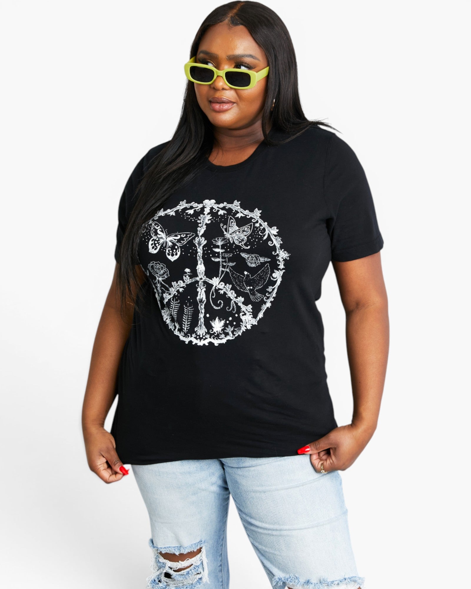 A woman modeling a black short-sleeve t-shirt with a white peace sign graphic filled with floral and butterfly designs, paired with light-wash distressed jeans and neon yellow sunglasses, against a plain white background.