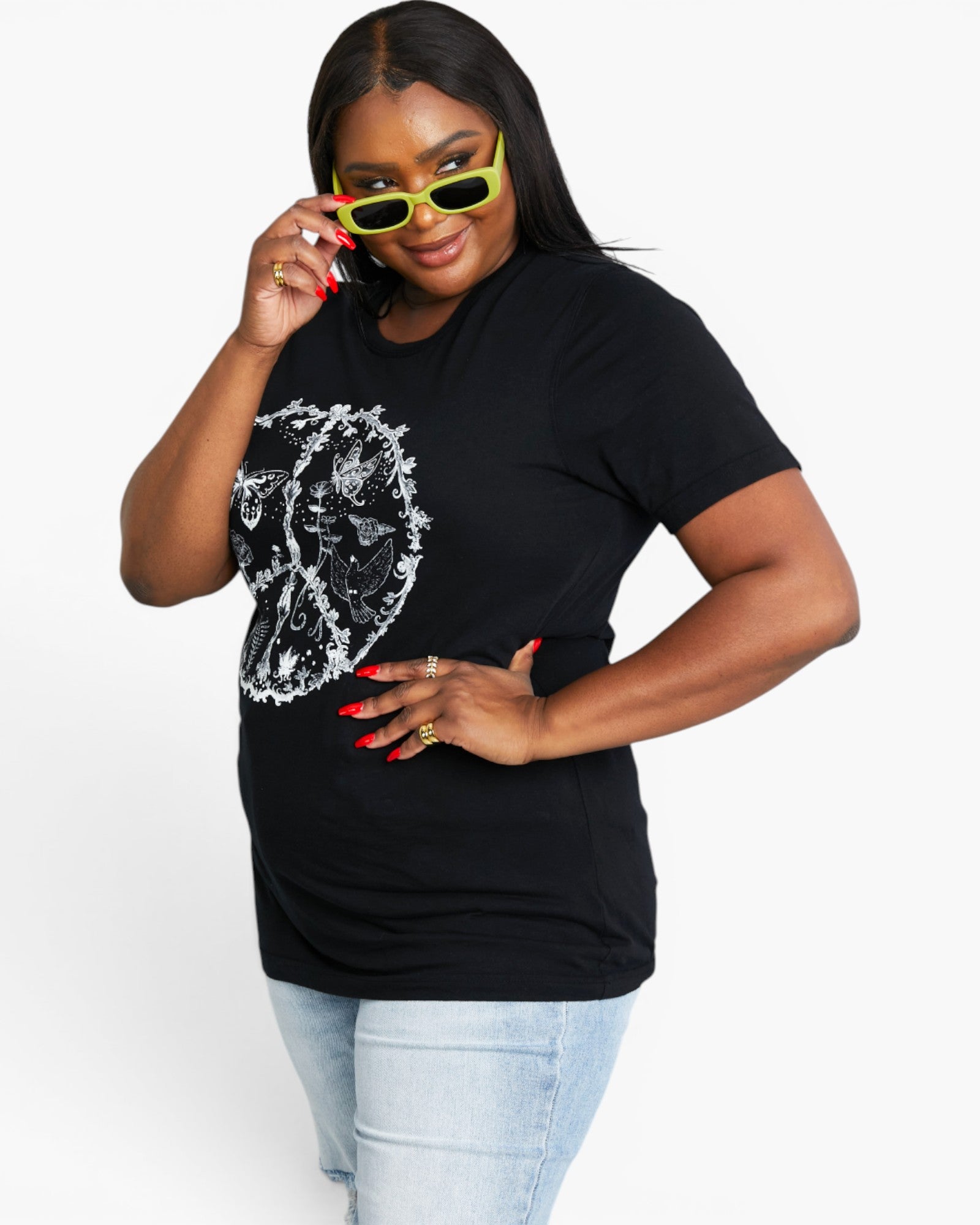 A woman modeling a black short-sleeve t-shirt with a white graphic print of a woman's face inside a peace sign. She is wearing light blue jeans, yellow sunglasses, and red nail polish against a plain white background.
