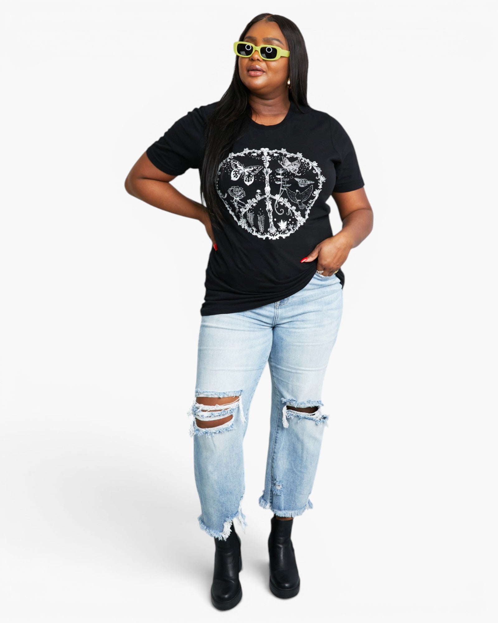 A woman modeling a black short-sleeve graphic t-shirt with a white skull-and-crossbones print, paired with light-wash distressed jeans and black ankle boots, against a plain white studio background.
