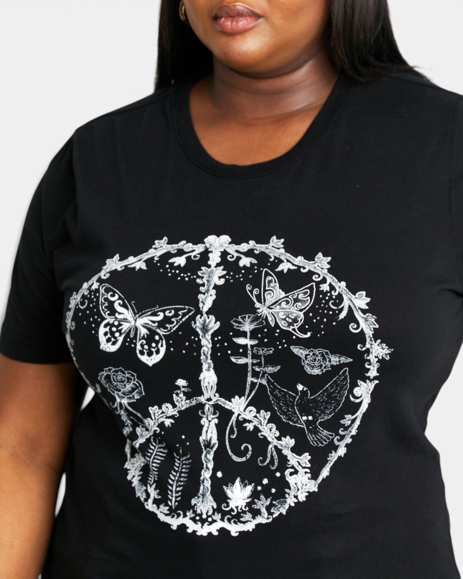 A plus-size black t-shirt featuring a white graphic print of a peace sign decorated with butterflies, flowers, and leaves. The shirt is modeled by a person against a plain light gray background.