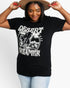 A woman wearing a black graphic t-shirt with a white distressed print that reads 'DESERT DREAMER' and 'DREAMER OF DESERT'. She is holding a wide-brimmed tan hat above her head and wearing light-wash blue jeans, set against a plain white studio background.