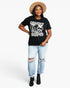 A woman modeling a black graphic t-shirt with a distressed white print that reads 'DESERT DREAMER', paired with light-wash ripped jeans, black ankle boots, and a tan wide-brim hat, against a plain white studio background.