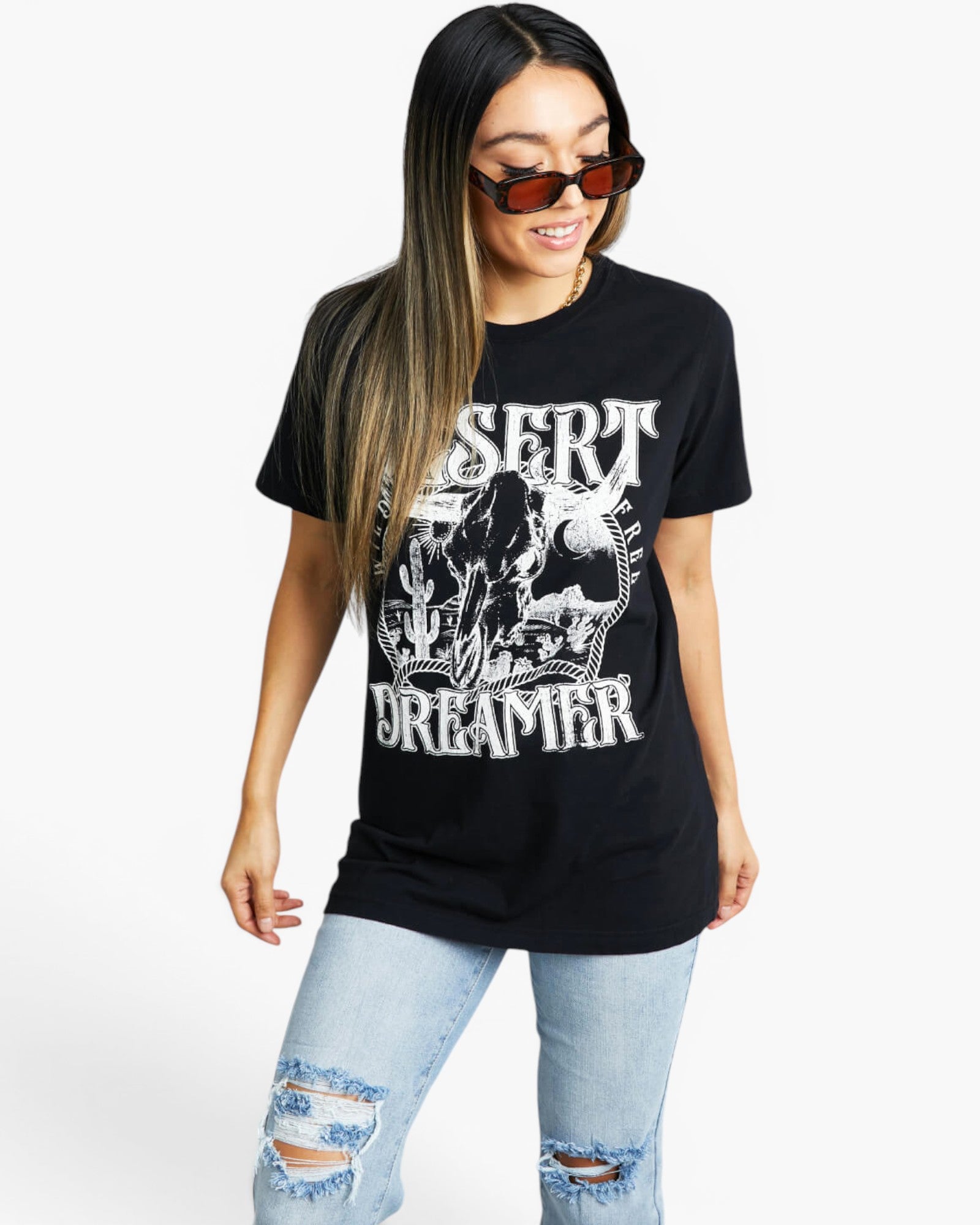 A woman modeling a black graphic t-shirt with a white distressed print that reads 'DESERT DREAMER' and features a skull design, paired with light-wash ripped jeans and sunglasses, against a plain white background.