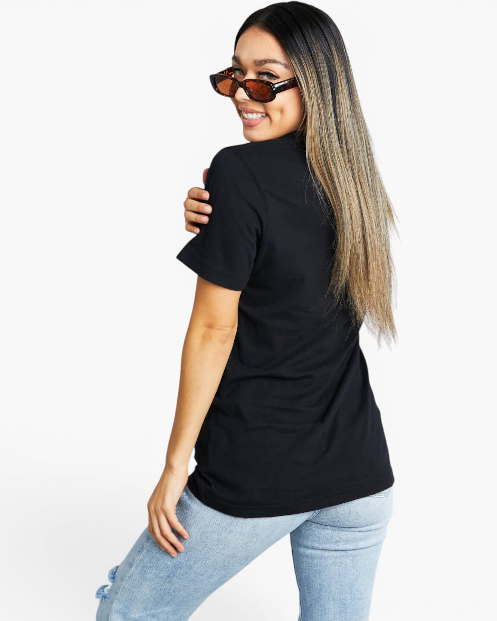 A woman with long blonde-highlighted hair and tortoiseshell sunglasses, wearing a black short-sleeve t-shirt and light-wash blue jeans, posing from a side-back angle against a plain white studio background.