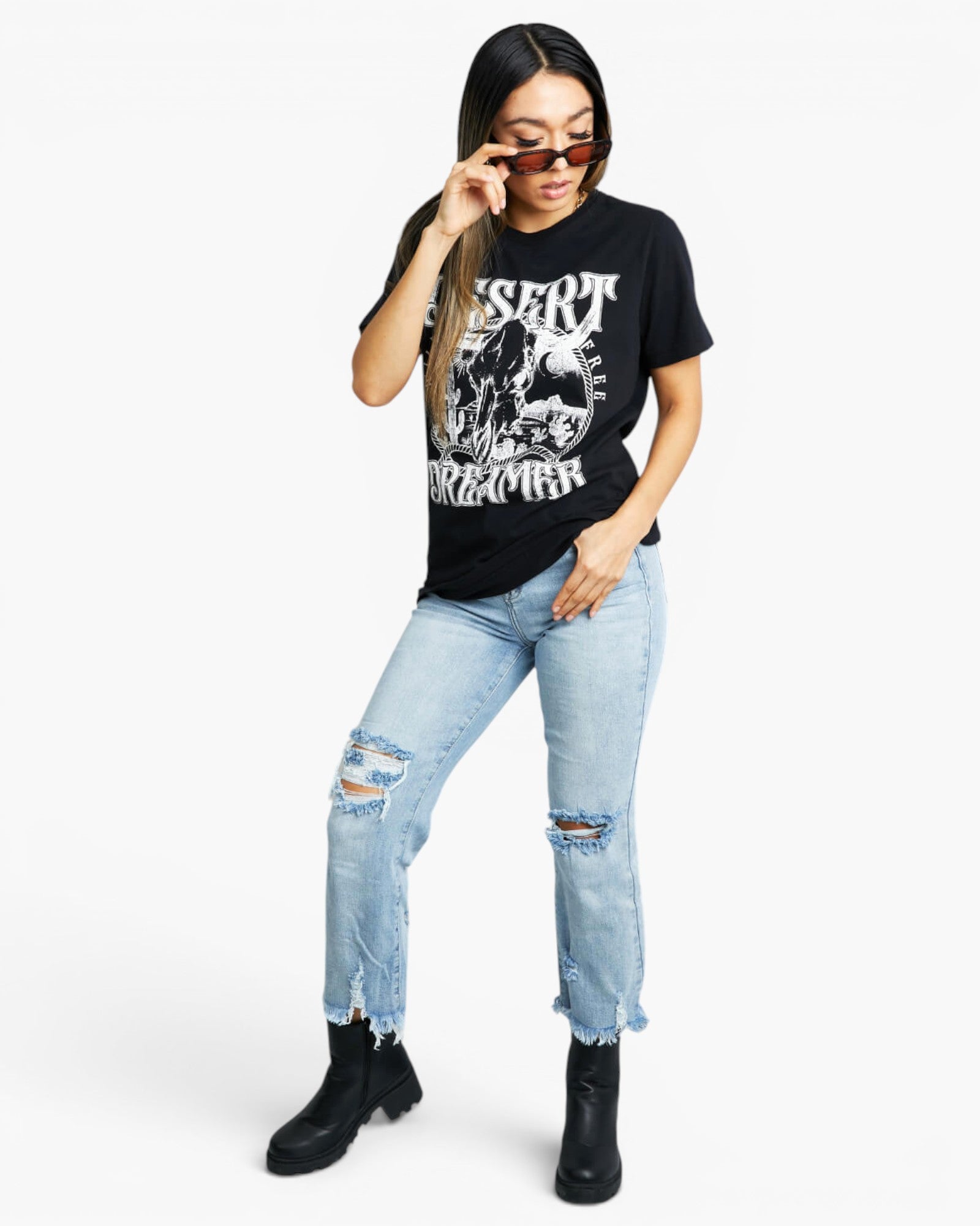 A woman modeling a black graphic t-shirt with a white skull print, paired with light-wash distressed jeans and black ankle boots, against a plain white background.
