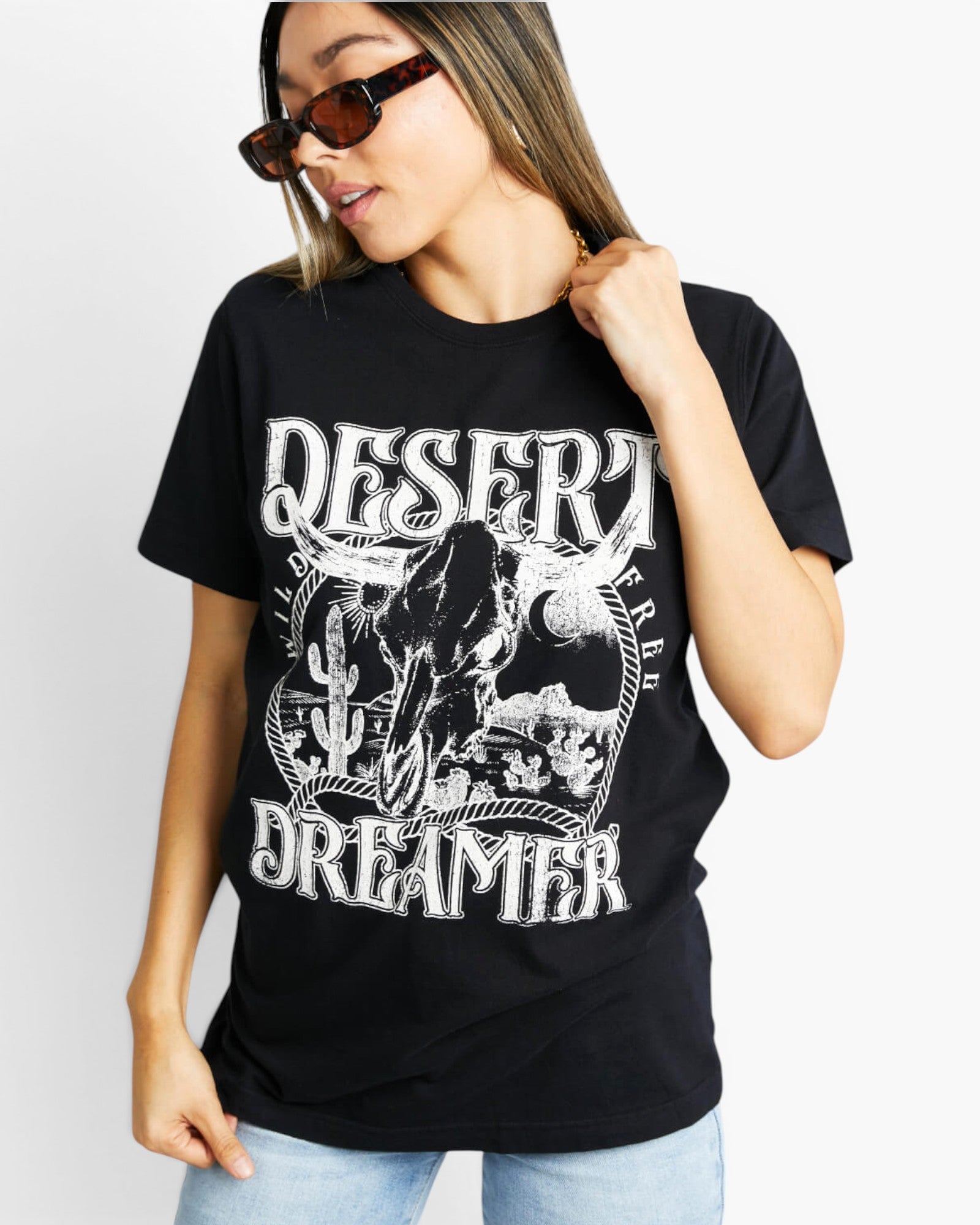A woman wearing a black graphic t-shirt with a white distressed print that reads 'DESERT DREAMER' and features a skull illustration, paired with light blue jeans against a plain white background.