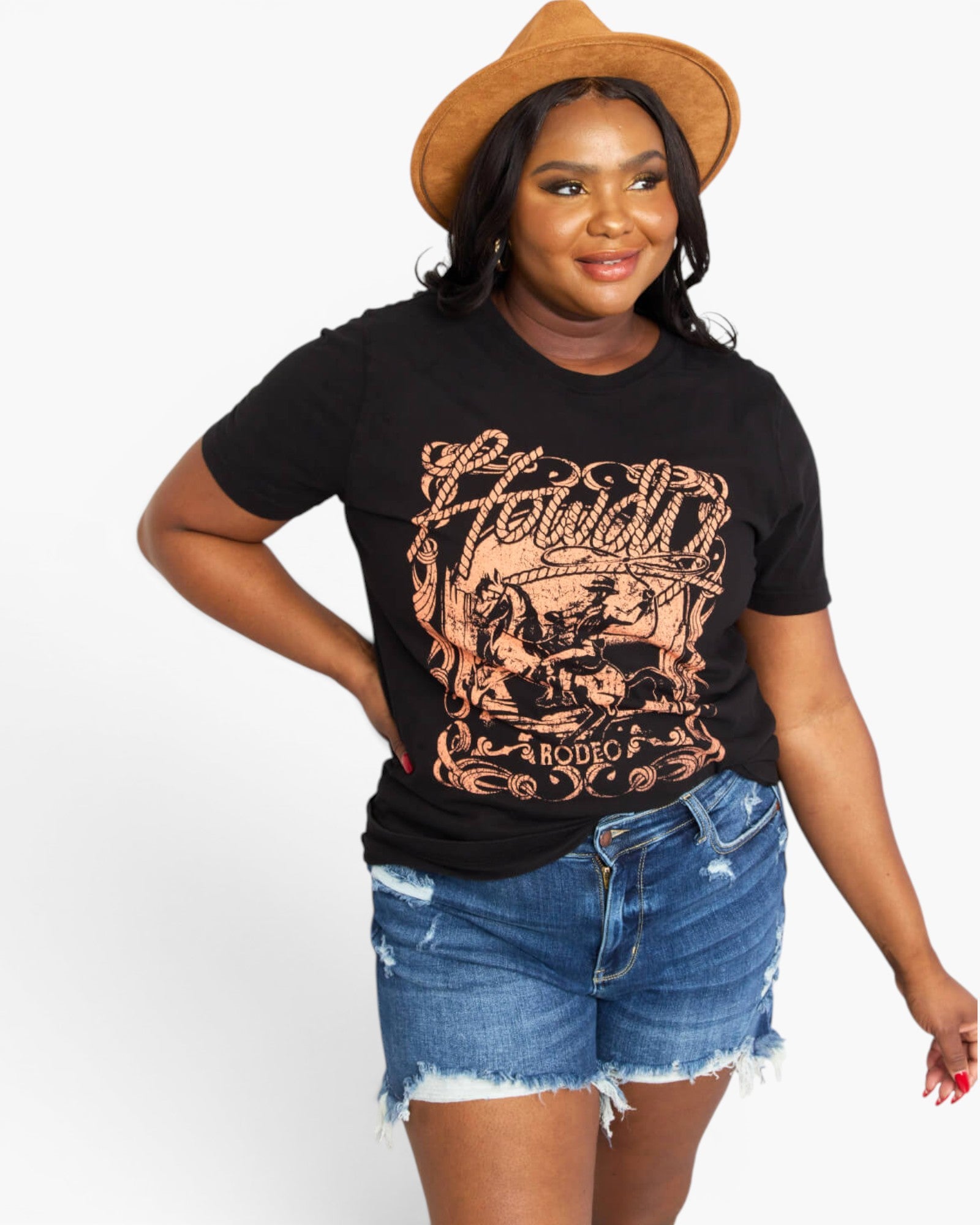 A woman modeling a black graphic t-shirt with a dusty rose graphic print that reads 'Howdy Rodeo', paired with blue denim shorts and a brown wide-brim hat, against a plain white background.