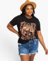 A woman modeling a black graphic t-shirt with a dusty rose graphic print that reads 'Howdy Rodeo', paired with blue denim shorts and a brown wide-brim hat, against a plain white background.