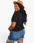 A woman wearing a black short-sleeve t-shirt, distressed denim shorts, and a brown wide-brim hat, photographed from the back against a plain white studio background.