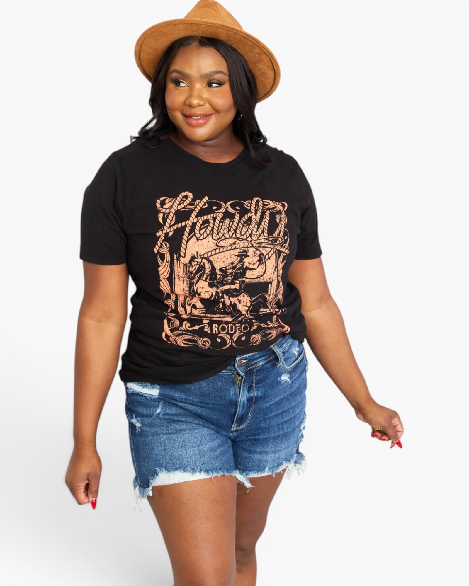 A woman modeling a black graphic t-shirt with a distressed rose gold design that reads 'Howdy Rodeo', paired with denim shorts and a brown hat, against a plain white background.