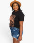 A woman wearing a black graphic t-shirt with a distressed copper-orange print, blue denim shorts, and a brown wide-brim hat, photographed against a solid white studio background.
