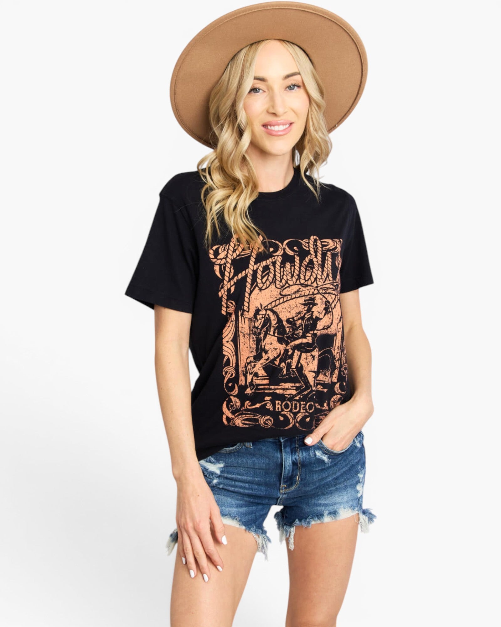A woman modeling a black graphic t-shirt with a distressed brown western-style print that reads 'Howdy Rodeo', paired with denim shorts and a tan wide-brim hat against a white studio background.