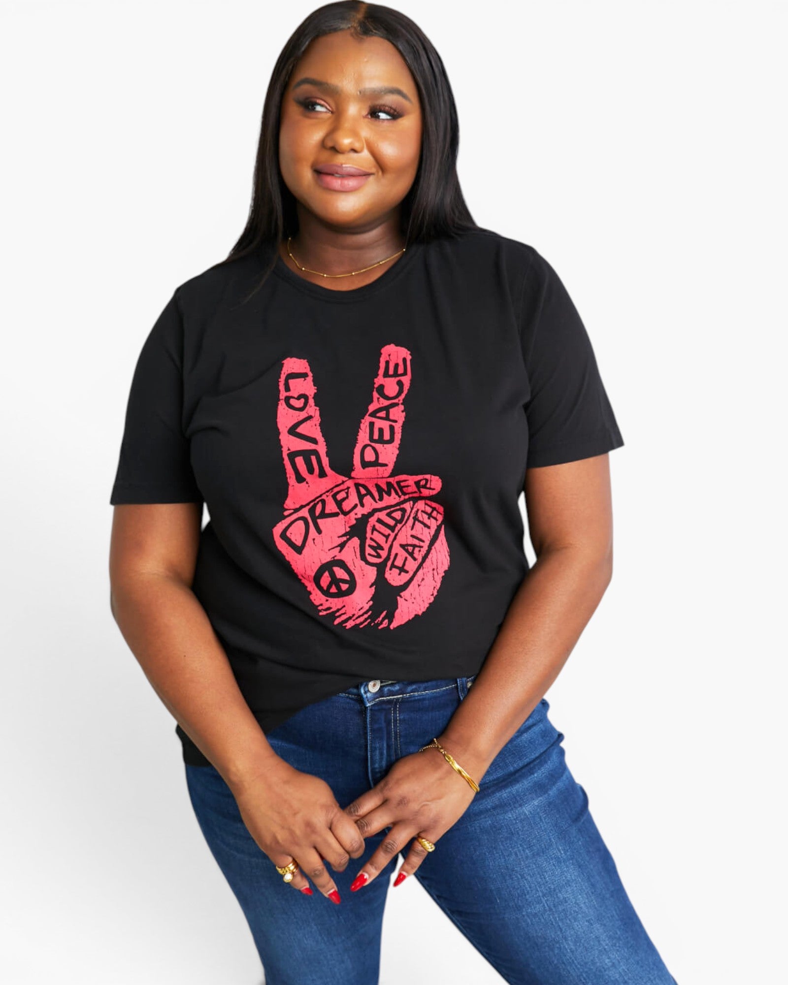 A woman wearing a black short-sleeve graphic T-shirt with a pink peace sign made from words including 'LOVE', 'PEACE', and 'DREAMER', paired with blue jeans, against a light gray studio background.