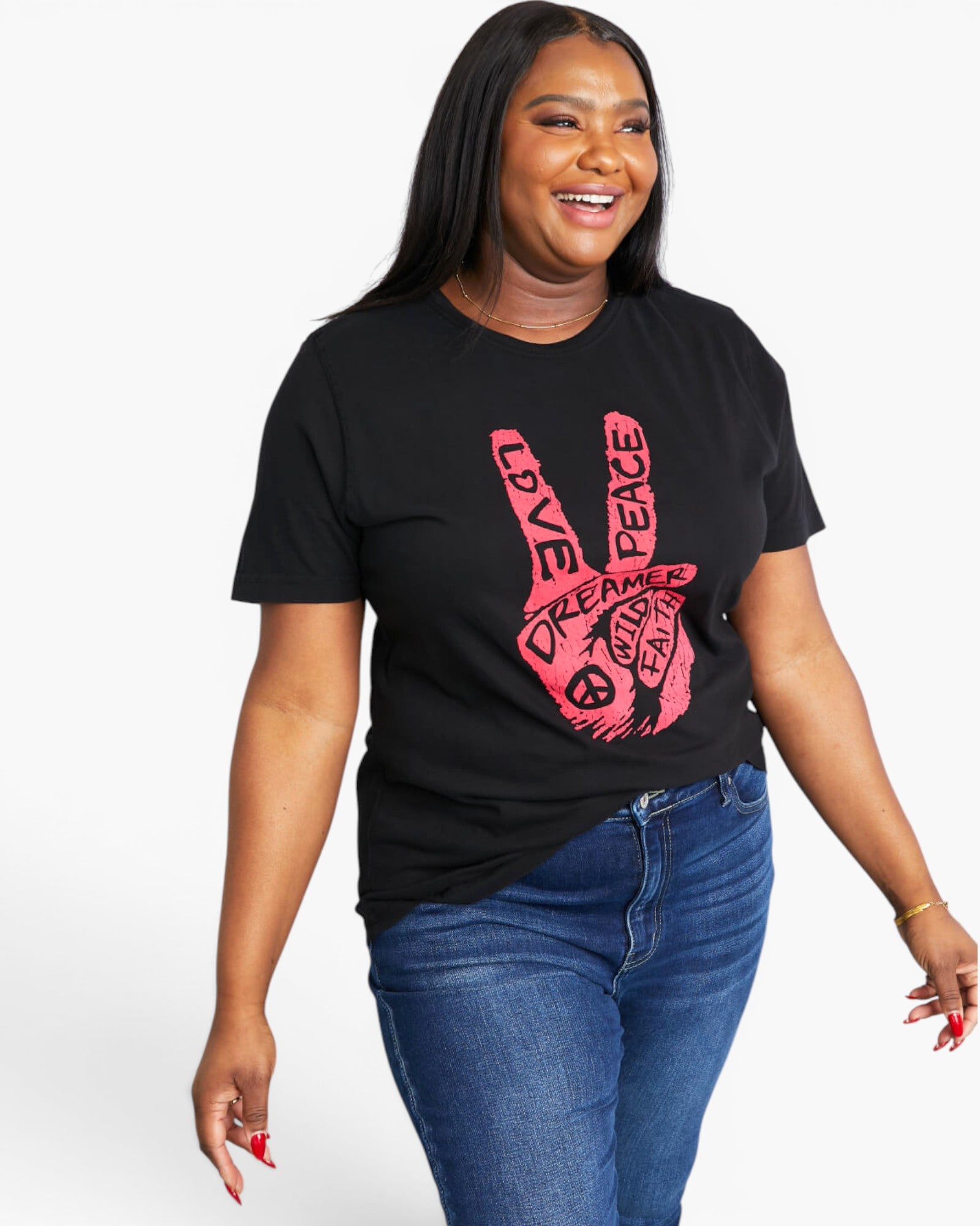 A woman modeling a black short-sleeve graphic t-shirt with a pink hand design containing the words 'LOVE', 'PEACE', and 'DREAMER'. She is wearing blue jeans against a plain white studio background.