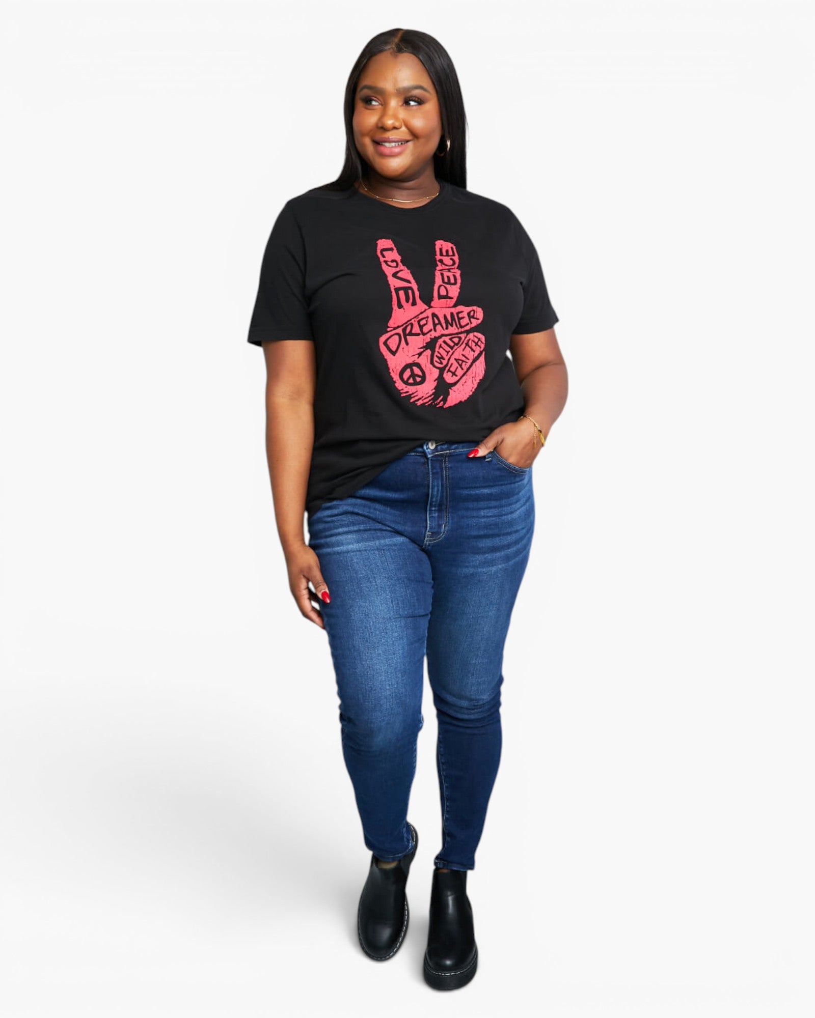 A woman modeling a black short-sleeve graphic t-shirt with a pink peace sign hand design, paired with blue skinny jeans and black ankle boots, against a light gray studio background.