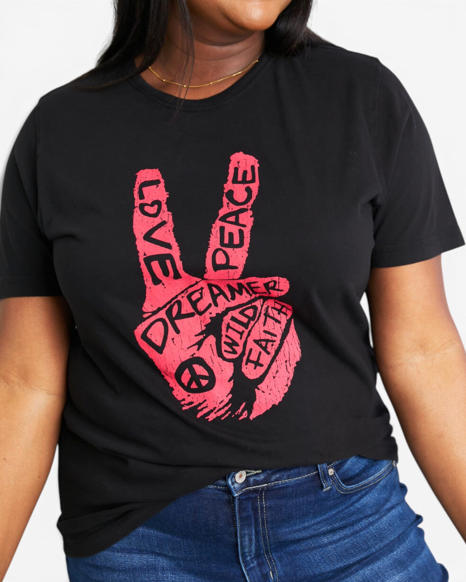 A woman wearing a black short-sleeve t-shirt with a pink graphic of a peace hand sign containing the words 'LOVE', 'PEACE', 'DREAMER', and 'WILD FAITH', paired with blue jeans.