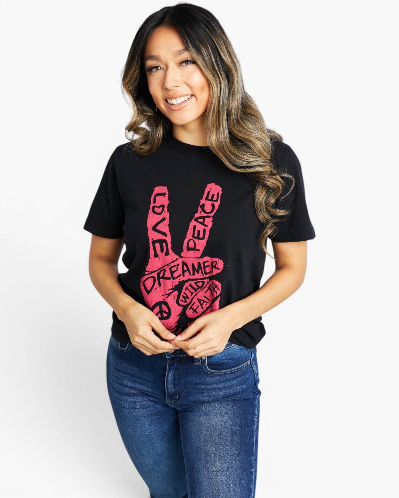 A woman wearing a black short-sleeve graphic t-shirt with a pink peace sign design containing the words 'LOVE', 'DREAMER', 'PEACE'. She is also wearing blue jeans, standing against a plain white studio background.