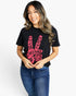 A woman wearing a black short-sleeve graphic t-shirt with a pink peace sign design containing the words 'LOVE', 'DREAMER', 'PEACE'. She is also wearing blue jeans, standing against a plain white studio background.