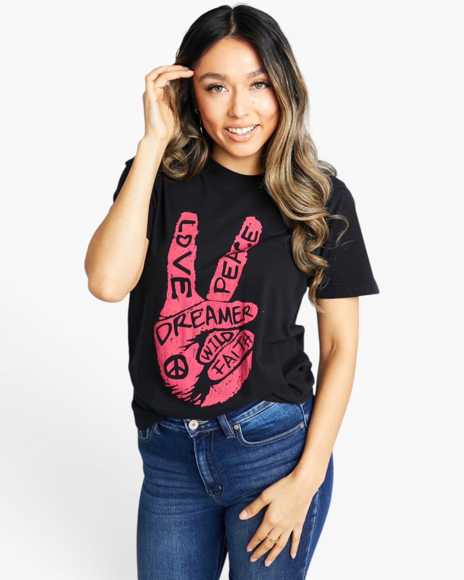 A woman wearing a black short-sleeve graphic t-shirt with a pink peace handprint design containing the words 'LOVE', 'PEACE', 'DREAMER', and 'WILD EARTH'. She is also wearing blue jeans, photographed against a solid light gray background.