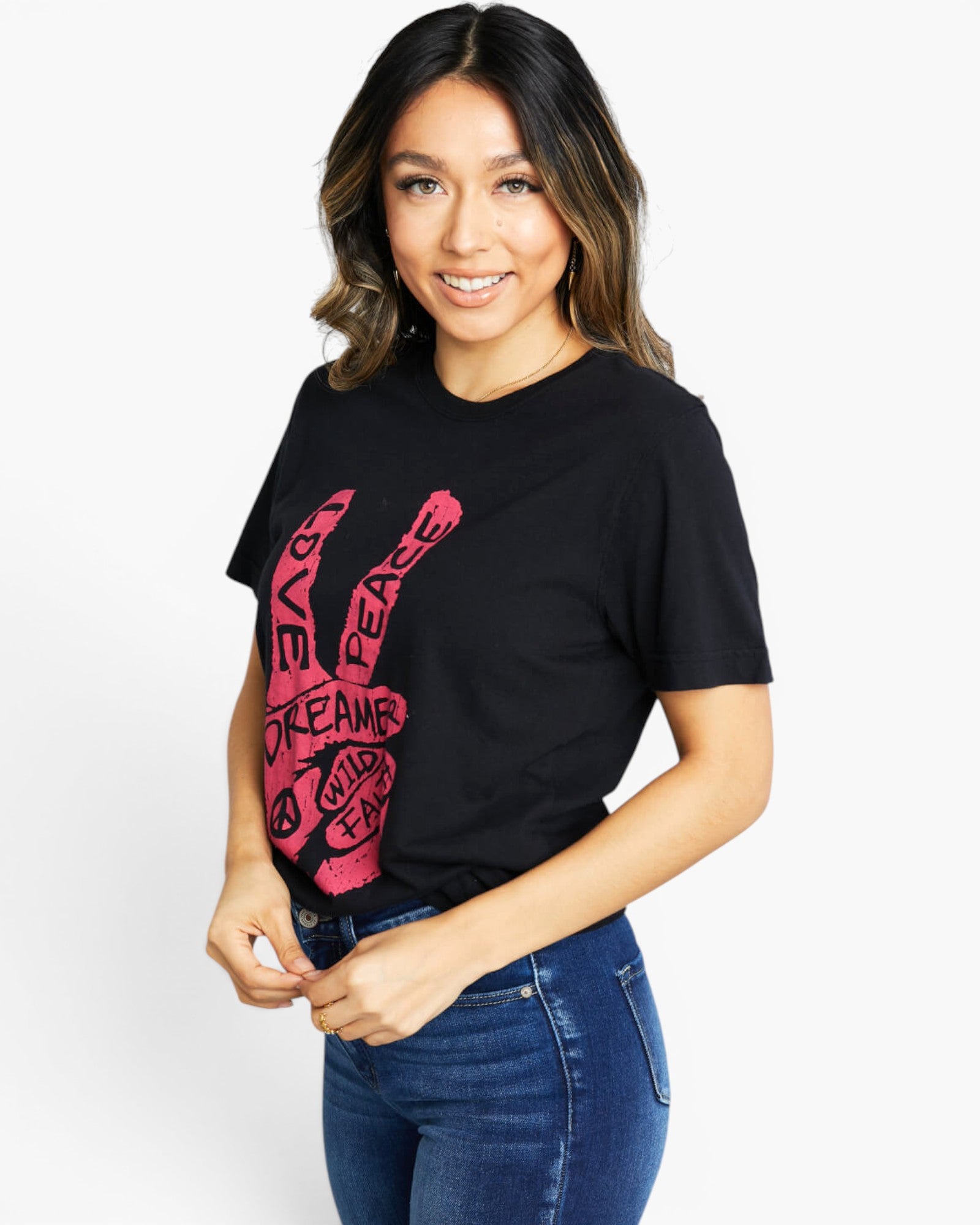 A woman wearing a black short-sleeve graphic t-shirt with a pink peace sign design and the words 'LOVE PEACE DREAMS' written inside the hand, paired with blue jeans, against a plain white background.