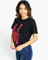 A woman wearing a black short-sleeve graphic t-shirt with a pink peace sign design and the words 'LOVE PEACE DREAMS' written inside the hand, paired with blue jeans, against a plain white background.