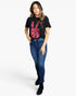 A woman modeling a black short-sleeve graphic T-shirt with a pink and red handprint design, paired with blue skinny jeans and black ankle boots, against a light gray studio background.