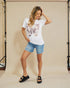 Woman wearing a white t-shirt with floral design and denim shorts against a beige backdrop