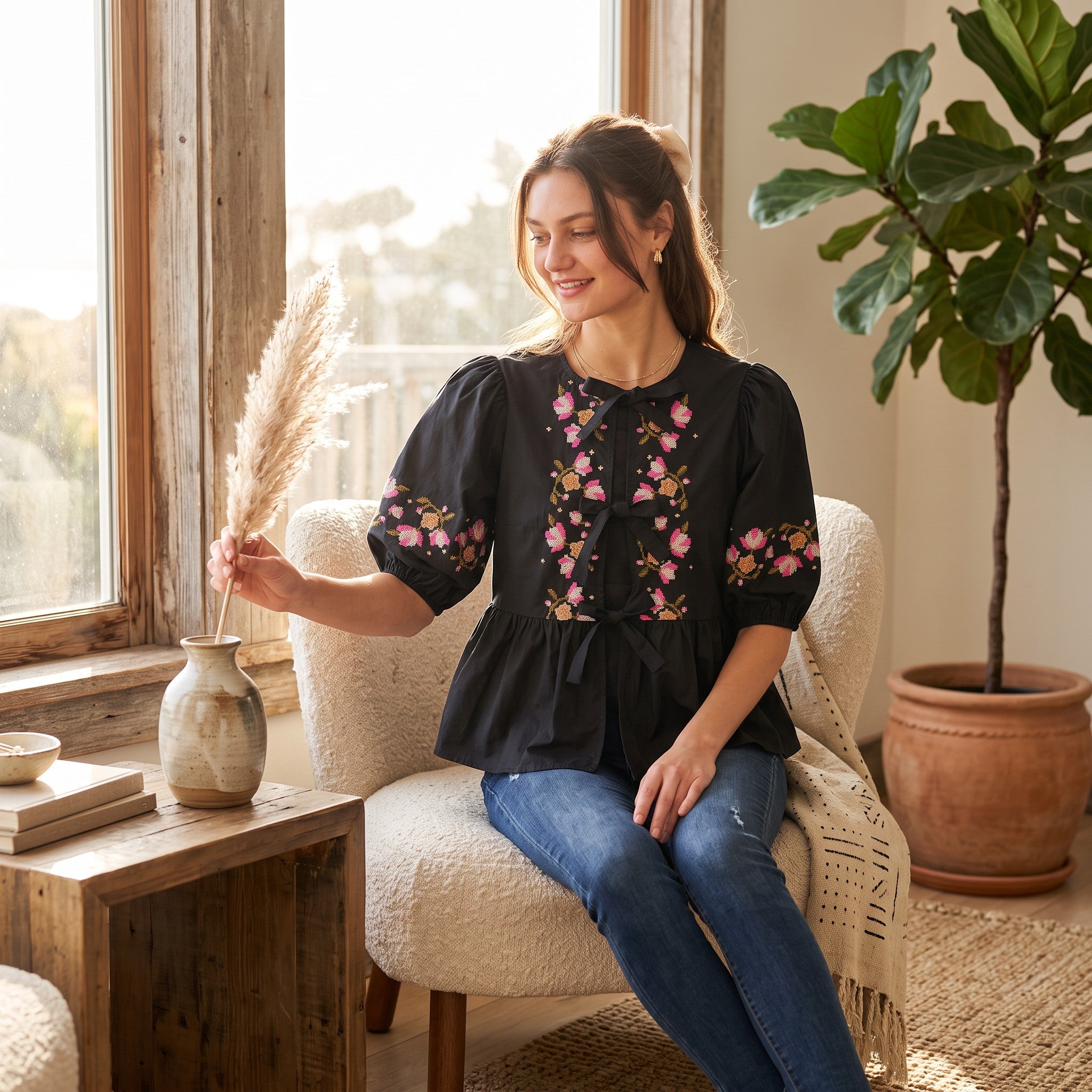 A woman sits in a cream boucle armchair by a window, wearing a black puff-sleeve blouse with pink and green floral embroidery on the bodice and sleeves, paired with blue jeans. She holds a dried pampas grass stem in a rustic ceramic vase on a wooden side table.