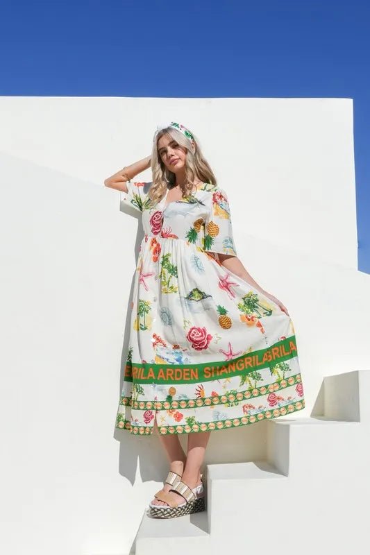 Woman wearing a colorful dress with floral patterns and text, standing against a white wall with blue sky.