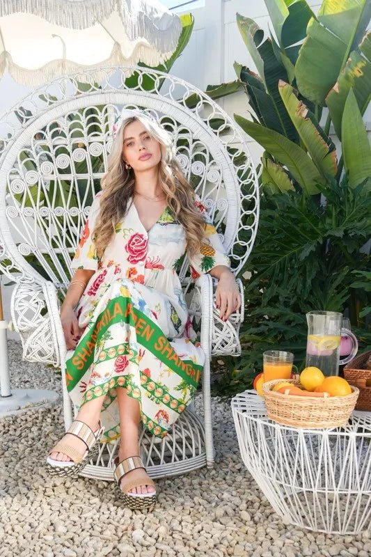 Woman in a floral dress sitting on a white wicker chair outdoors with plants and a table in the background.