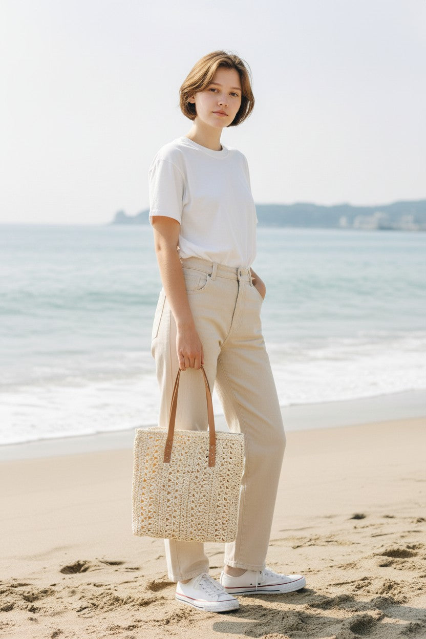 Woman standing on a beach wearing a white shirt, beige pants, and white sneakers, holding a woven bag.